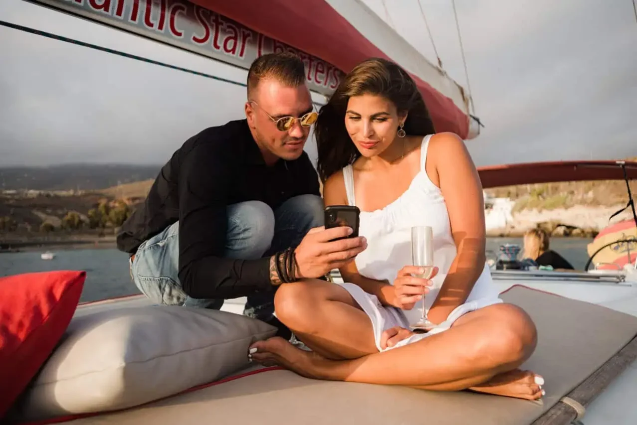 A couple relaxes on a boat, closely reviewing something on a smartphone, with the "Atlantic Star" logo visible, showcasing a serene moment during a Tenerife boat trip.